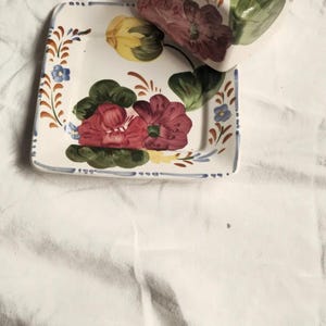 May include: A square ceramic dish and lid set with a floral design. The set features a white base with hand-painted red, yellow, and green flowers. The edges are trimmed with blue and brown accents.