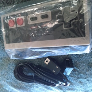May include: A black and gray retro video game controller with red buttons, packaged in a clear plastic bag. A black cable and connector are visible below the controller, suggesting it's a wired device.