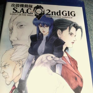 May include: A blue Blu-ray case for Ghost in the Shell S.A.C. 2nd GIG Stand Alone Complex 4-Disc Complete Collection. The cover features an anime illustration of four characters and a white bird, with the title text printed in black at the top.