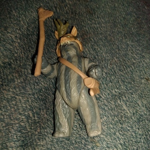 May include: A Star Wars Ewok action figure. The figure is gray and tan, with a tan satchel and a staff. The Ewok is wearing a helmet with a green leaf design. The figure is standing on a textured surface.