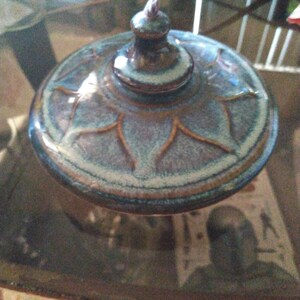 May include: A round ceramic lid with a central knob handle, featuring a glazed finish in shades of blue, purple, and brown. The surface is decorated with a raised, star-like radial pattern that extends from the center toward the outer edge.