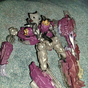 May include: A purple, gray, and gold action figure in a robotic design. The figure has multiple articulated joints, with a complex arrangement of panels and mechanical details. The figure is posed on a textured surface.