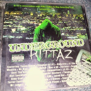 May include: A CD case with the title "UNDERGROUND KITTAZ" in green and white lettering. The cover features a hooded figure in green against a cityscape background. The Parental Advisory label is visible.