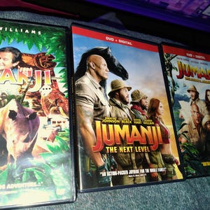 May include: Three DVD cases of the movie "Jumanji" are displayed. The first features Robin Williams, the second "Jumanji: The Next Level" with Dwayne Johnson, and the third with the cast. The cases have colorful graphics and the title "Jumanji".