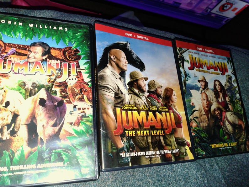 May include: Three DVD cases of the movie "Jumanji" are displayed. The first features Robin Williams, the second "Jumanji: The Next Level" with Dwayne Johnson, and the third with the cast. The cases have colorful graphics and the title "Jumanji".