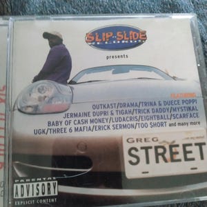 May include: CD case for Slip-N-Slide Records featuring a man in a purple shirt and a car graphic. The CD case features the text "GREG STREET" on a license plate and a parental advisory warning.