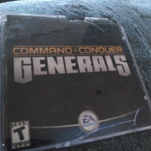 May include: A damaged black video game case for "Command & Conquer Generals". The case has a large crack across the front and features the EA Games logo and a "T" for Teen rating.