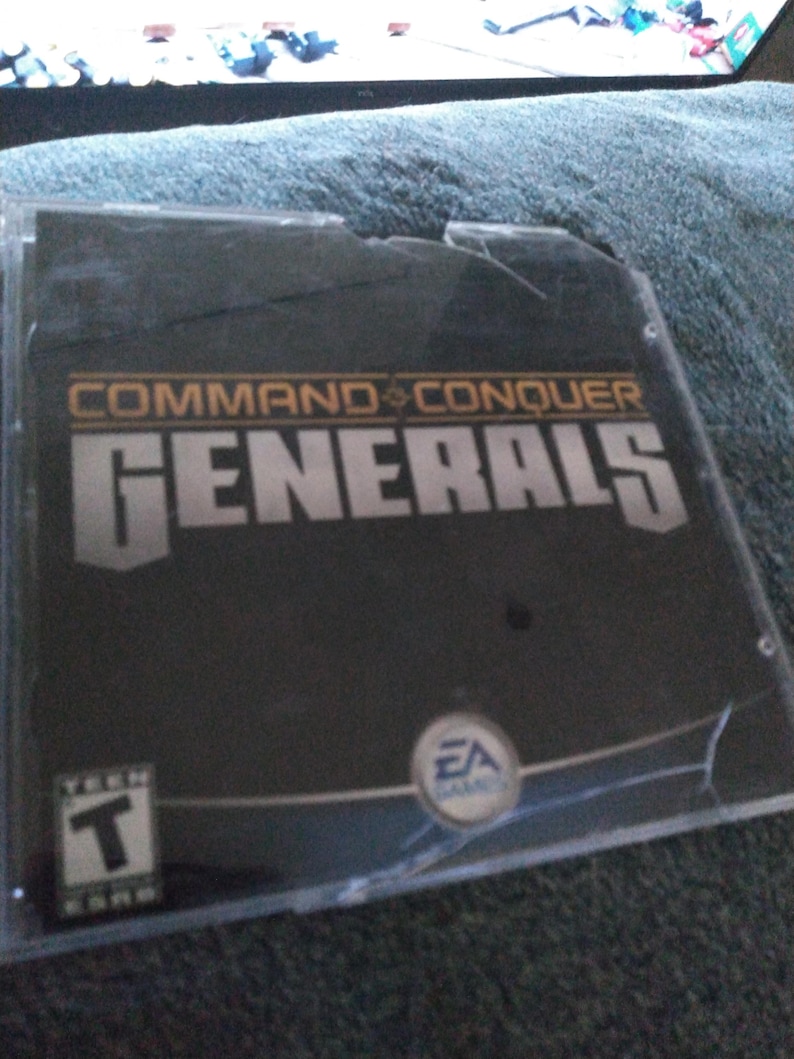 May include: A cracked black video game case for "Command & Conquer Generals". The case has a large crack across the front and features the EA Games logo and a "T" for Teen rating.