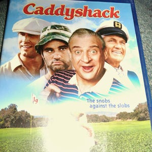 May include: Blu-ray disc of the movie "Caddyshack" with a blue case. The cover features four men, the title in red, and the tagline "The snobs against the slobs."
