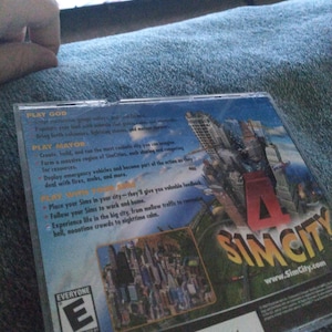 May include: A SimCity PC game case. The cover shows a cityscape with the game's logo. The back of the case has text describing gameplay features, including playing as God or Mayor. The ESRB rating is visible.