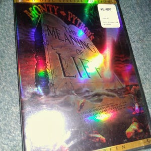 May include: A two-disc special edition DVD of Monty Python's The Meaning of Life. The cover features a stone tablet graphic, iridescent rainbow light reflections, and a white retail sticker. The text lists the writers and stars of the film.