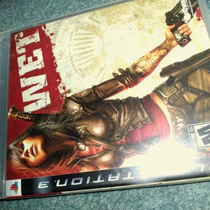 May include: A PlayStation 3 video game case for the game WET. The cover art features a character with long dark hair, a brown leather jacket, and tattoos, holding a black handgun. The title WET is in large white letters on a red background.