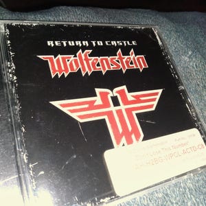 May include: A CD case for the video game "Return to Castle Wolfenstein." The cover is black with red text and a red emblem. The text "Wolfenstein" is in a stylized font. A white sticker with text is on the lower right.