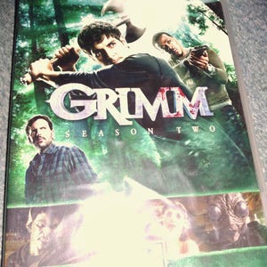 May include: A DVD case for Grimm Season Two featuring a dark forest background with three main characters. One person holds an axe, another holds a handgun, and a third stands below. The bottom panel displays three distinct creature-like faces.