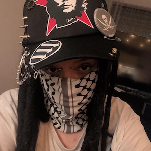 Könnte beinhalten: Schwarze Baseballkappe mit rotem Stern und Che Guevara-Grafik. Die Kappe hat Metallspitzen, einen Piraten-Totenkopf-Aufnäher und andere dekorative Elemente. Ein weißes und schwarzes gemustertes Gesichtstuch ist ebenfalls sichtbar.