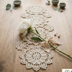 Puede incluir: Camino de mesa de ganchillo color crema con un diseño floral, adornado con una peonía blanca y una cinta delicada. El camino de mesa está colocado sobre una mesa de madera, junto a pequeños cuencos y pendientes.
