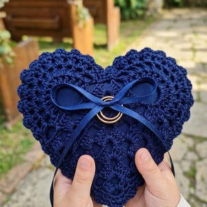 Puede incluir: Un cojín para anillos en forma de corazón azul marino con un diseño de ganchillo. Una cinta azul marino está atada alrededor del centro, sosteniendo dos anillos de boda dorados. El cojín es sostenido por dos manos.