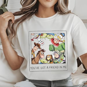 Puede incluir: Camiseta blanca con un diseño enmarcado de personajes de Toy Story, incluyendo a Woody, Buzz Lightyear y Rex. El diseño está rodeado por un borde negro. El texto "YOU'VE GOT A FRIEND IN ME" está debajo.