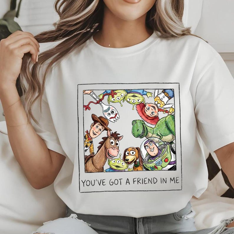 May include: White t-shirt featuring a framed graphic of characters from the animated film Toy Story. The graphic includes Woody, Buzz Lightyear, Rex, and other characters. The text "YOU'VE GOT A FRIEND IN ME" is below the graphic.