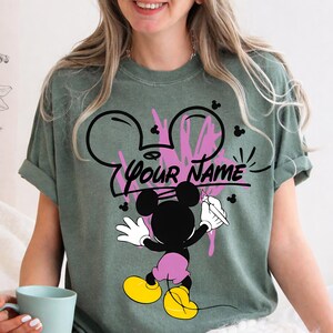May include: A sage green short-sleeve t-shirt featuring a graphic of a cartoon mouse character holding a paintbrush. The design includes a pink paint splatter effect, a black outline of a mouse head, and the black text "YOUR NAME" in the center.