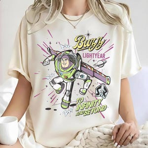 May include: A cream-colored t-shirt featuring a graphic of Buzz Lightyear in a space suit with purple, green, and white accents. The text reads Buzz Lightyear and To Infinity and Beyond, accompanied by starbursts, a planet, and a rocket ship emblem.