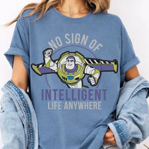 May include: A blue t-shirt featuring a graphic of the character Buzz Lightyear with his wings extended. The text above and below the character reads, "NO SIGN OF INTELLIGENT LIFE ANYWHERE" in a bold, sans-serif font, centered on the chest of the shirt.