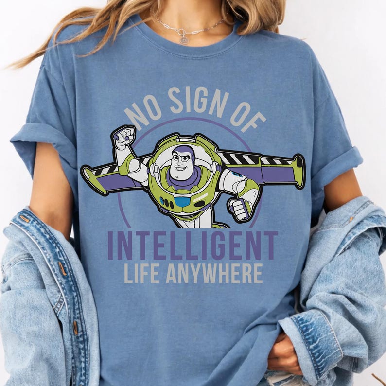 May include: A blue t-shirt featuring a graphic of the character Buzz Lightyear with his wings extended. The text above and below the character reads, "NO SIGN OF INTELLIGENT LIFE ANYWHERE" in a bold, sans-serif font, centered on the chest of the shirt.