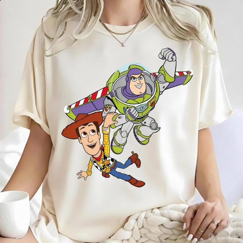 May include: A cream-colored short-sleeve t-shirt featuring a graphic print of the characters Woody and Buzz Lightyear from Toy Story. Woody is wearing his signature cowboy hat and outfit, while Buzz Lightyear is depicted flying with wings extended.