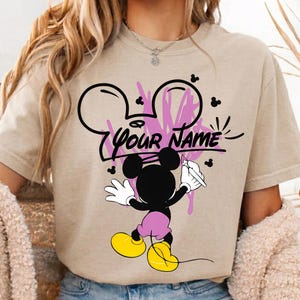 May include: A beige short-sleeved t-shirt featuring a graphic of a black Mickey Mouse character holding a paintbrush. The design includes pink paint splatters, small black mouse ear silhouettes, and the black text "YOUR NAME" in the center.
