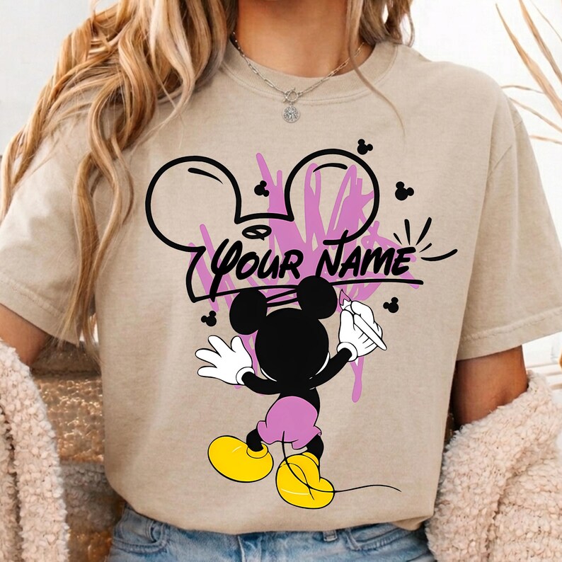 May include: A beige short-sleeved t-shirt featuring a graphic of a black Mickey Mouse character holding a paintbrush. The design includes pink paint splatters, small black mouse ear silhouettes, and the black text "YOUR NAME" in the center.