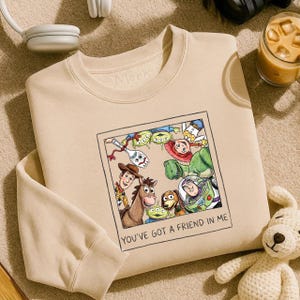 May include: Beige crewneck sweatshirt featuring a framed graphic of Toy Story characters, including Woody, Buzz Lightyear, and the aliens. The text "YOU'VE GOT A FRIEND IN ME" is below the image. A white stuffed dog is in the bottom right corner.