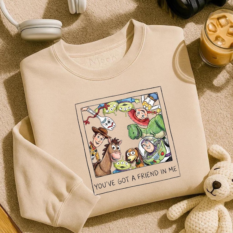 May include: Beige crewneck sweatshirt featuring a framed graphic of Toy Story characters, including Woody, Buzz Lightyear, and the aliens. The text "YOU'VE GOT A FRIEND IN ME" is below the image. A white stuffed dog is in the bottom right corner.