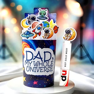 May include: A cylindrical container with a space theme, featuring the text "DAD, MY WHOLE UNIVERSE." It is decorated with stickers of astronauts, planets, rockets, and aliens. A bookmark with an astronaut on a crescent moon is also visible.