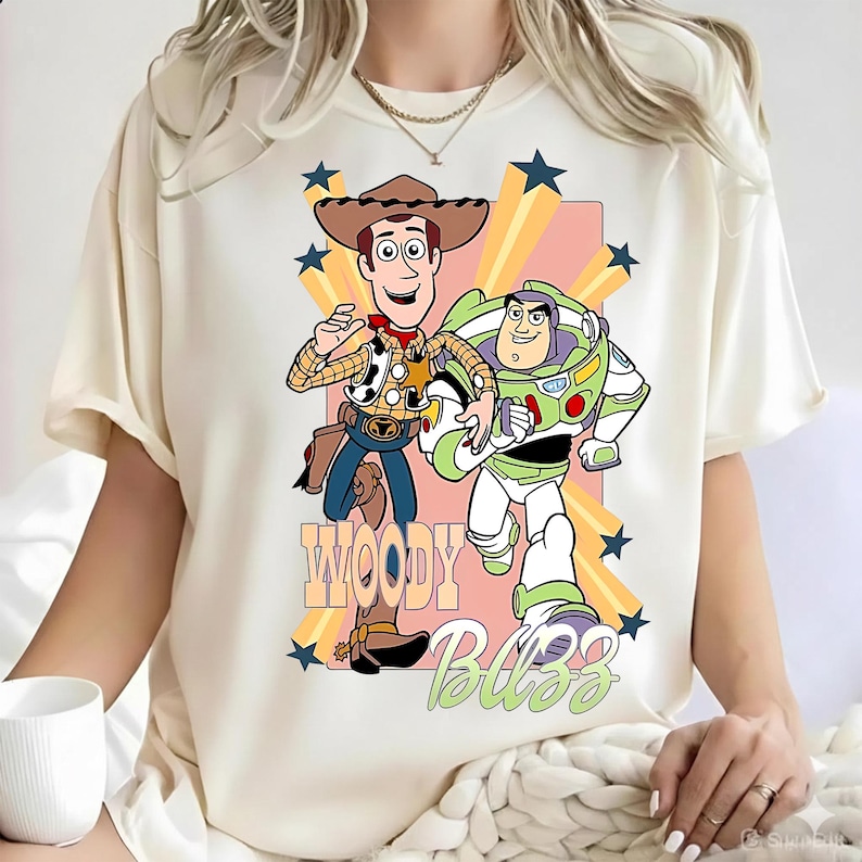 May include: A cream-colored short-sleeve t-shirt featuring a graphic print of Woody and Buzz Lightyear from Toy Story. The design includes yellow star accents, a pink rectangular background, and the text Woody and Buzz in stylized lettering.