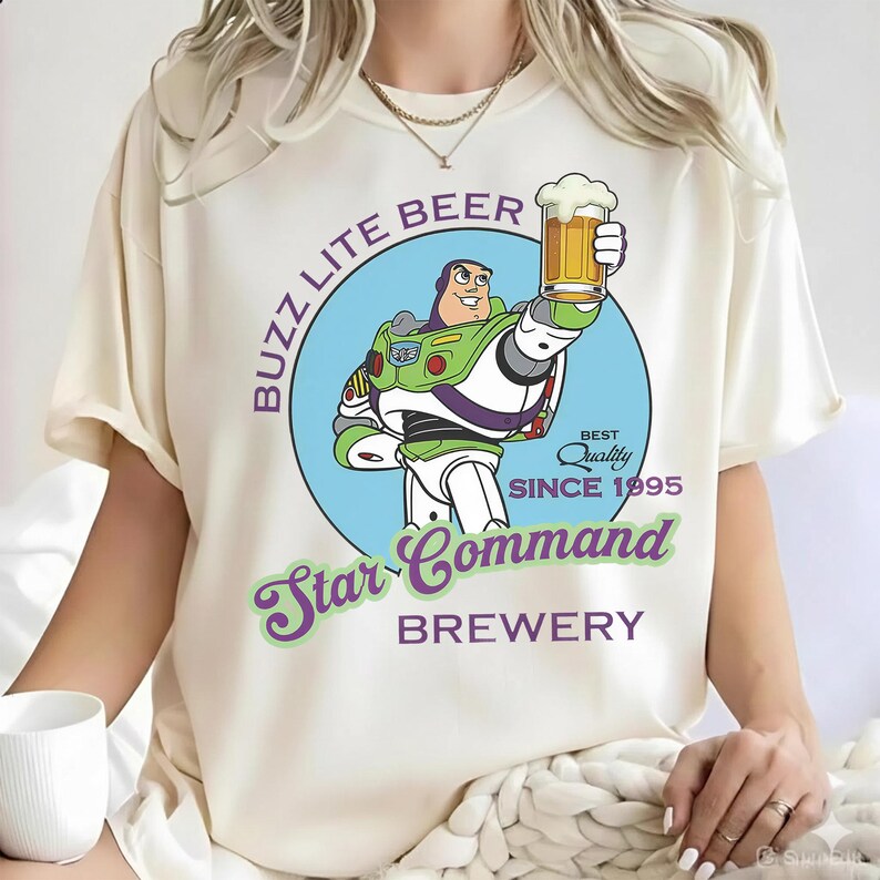 May include: A cream-colored t-shirt featuring a graphic of a space ranger character holding a beer mug. The text reads BUZZ LITE BEER, BEST QUALITY SINCE 1995, Star Command, and BREWERY. The design is set within a light blue circle on the front.