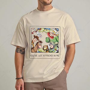 May include: Beige t-shirt featuring a framed illustration of characters from the animated film Toy Story. The image includes Woody, Buzz Lightyear, and other characters. The text "YOU'VE GOT A FRIEND IN ME" is printed below the illustration.
