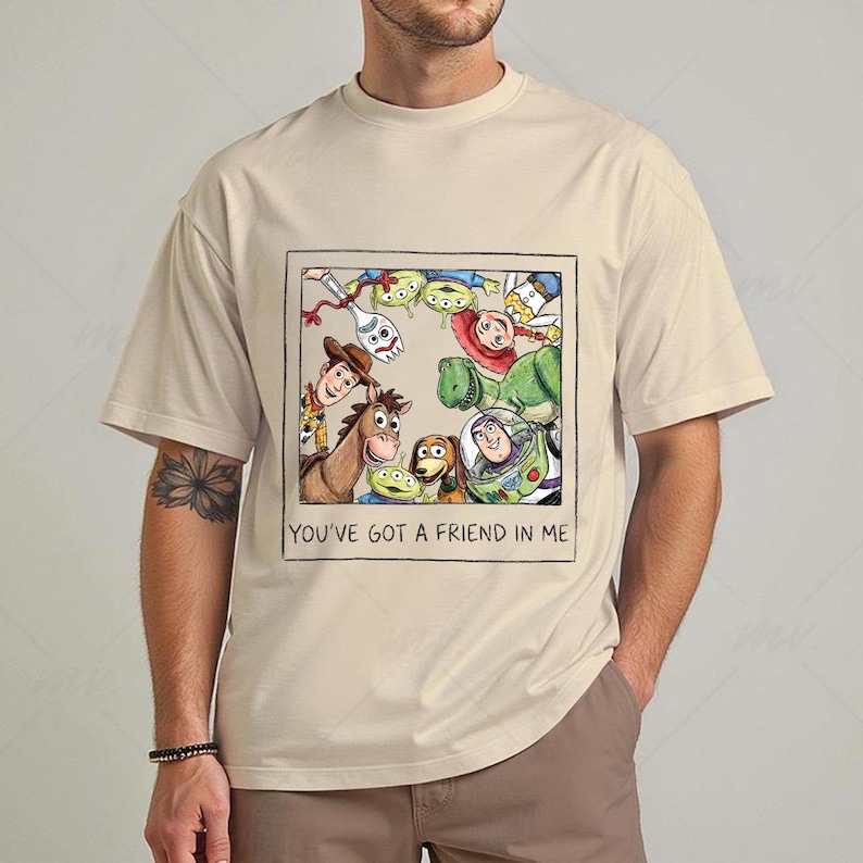 May include: Beige t-shirt featuring a framed illustration of characters from the animated film Toy Story. The image includes Woody, Buzz Lightyear, and other characters. The text "YOU'VE GOT A FRIEND IN ME" is printed below the illustration.