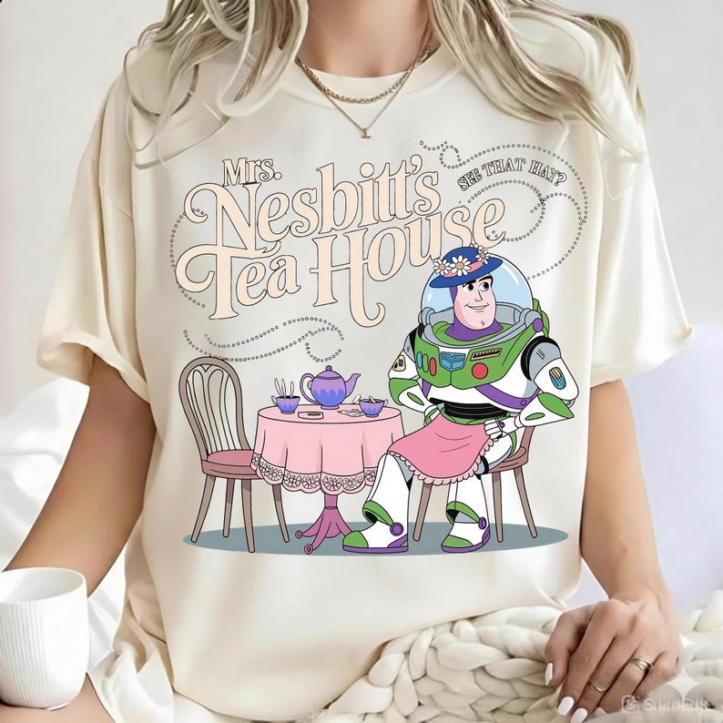 May include: A cream-colored t-shirt featuring a graphic of a space ranger character wearing a floral hat and pink apron, sitting at a table with a teapot and cups. The text reads Mrs. Nesbitt's Tea House and See That Hat? in a retro font.