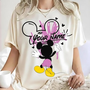 May include: A cream-colored t-shirt featuring a graphic of a black mouse character with yellow shoes and pink shorts, holding a pen. Above the character is a pink scribbled design and the black text "YOUR NAME" written in a stylized font.