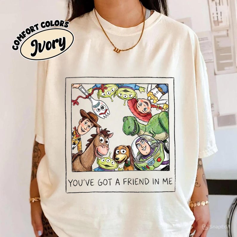May include: Ivory-colored t-shirt featuring a framed illustration of characters from a popular animated movie. The illustration includes a cowboy, a horse, a dinosaur, and other characters. The text "YOU'VE GOT A FRIEND IN ME" is below the illustration.