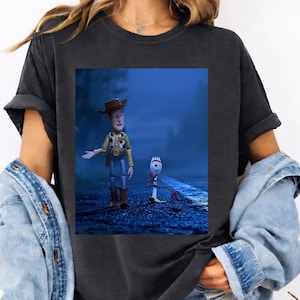 May include: A charcoal gray t-shirt featuring a graphic print of the characters Woody and Forky from Toy Story standing on a dark, blue-toned road at night. The shirt has short sleeves and a crew neckline, worn with a blue denim jacket.