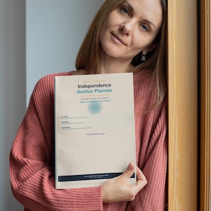 May include: A woman in a pink sweater holds a planner titled "Independence Anchor Planner." The planner has a light blue and white cover with text and a circular design. The image is taken indoors, near a wooden door frame.