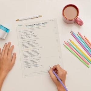 Könnte beinhalten: Draufsicht auf einen Arbeitsplatz mit einer Checkliste mit dem Titel "Documents & Reality Checklist". Eine Hand schreibt mit einem lila Bleistift auf die Checkliste. Weitere Artikel sind eine Kaffeetasse, Buntstifte, ein Stift und eine Rolle Klebeband.