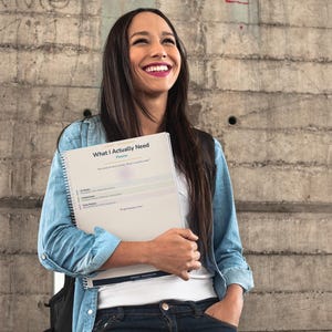 May include: A spiral-bound planner titled "What I Actually Need" is held by a person. The planner has a white cover with text and a light purple accent. The person is wearing a denim shirt and jeans, standing in front of a concrete wall.
