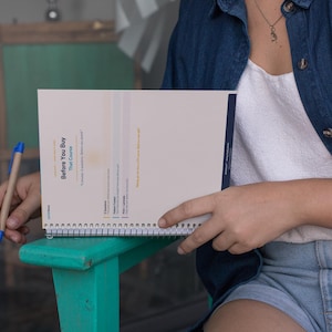 May include: A spiral-bound notebook with the text "Before You Buy That Course" is held open. A blue and tan pen rests nearby. The notebook is on a teal bench. The person is wearing a denim shirt and jean shorts.