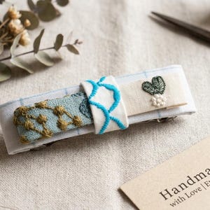 以下が含まれることがあります： 手作りのヘアクリップ。刺繍が施されており、水色と白のチェック柄、青色の刺繍デザイン、緑の葉のデザインが特徴です。「Handmade with Love | Etsy Shop」と書かれたタグも表示されています。