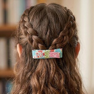 以下が含まれることがあります： 青、ピンク、赤、緑の色合いで様々な花柄が施されたパッチワークデザインの長方形の布製ヘアクリップ。このヘアアクセサリーは後頭部に留められており、ウェーブのかかった茶色の髪の三つ編み部分を2つまとめています。