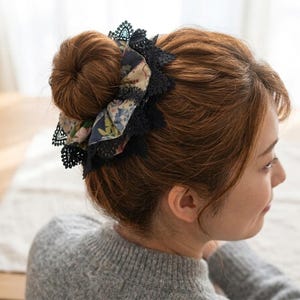 以下が含まれることがあります： 黒いレースのトリムが付いた花柄のヘアアクセサリーが、お団子ヘアに着用されています。アクセサリーは、青、ピンク、クリーム色の混合色です。髪は茶色で、その人は灰色のセーターを着ています。