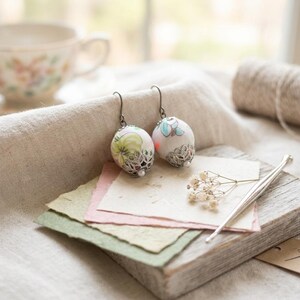 May include: A pair of drop earrings featuring round white ceramic beads with delicate floral patterns in green, blue, and pink. The beads are accented with intricate silver-toned metal filigree caps at the base and hang from simple metal ear wires.