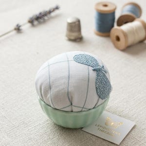 以下が含まれることがあります： 白と青のチェック柄のピンクッション。青い刺繍のデザインが施され、淡い緑色のセラミックボウルに入っています。糸巻きや指ぬきなど、裁縫道具が近くにあります。「LEPIDOPTERA THREADS」と書かれたタグが付いています。